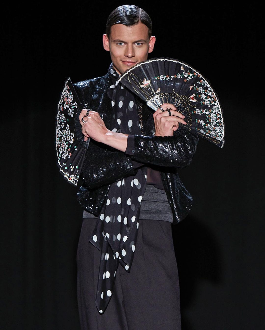 Male fashion model poses in a sequined jacket, black pants, and polka-dot scarf while holding a decorative fan against a black background.