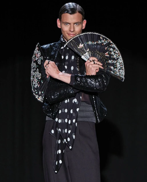 Male fashion model poses in a sequined jacket, black pants, and polka-dot scarf while holding a decorative fan against a black background.