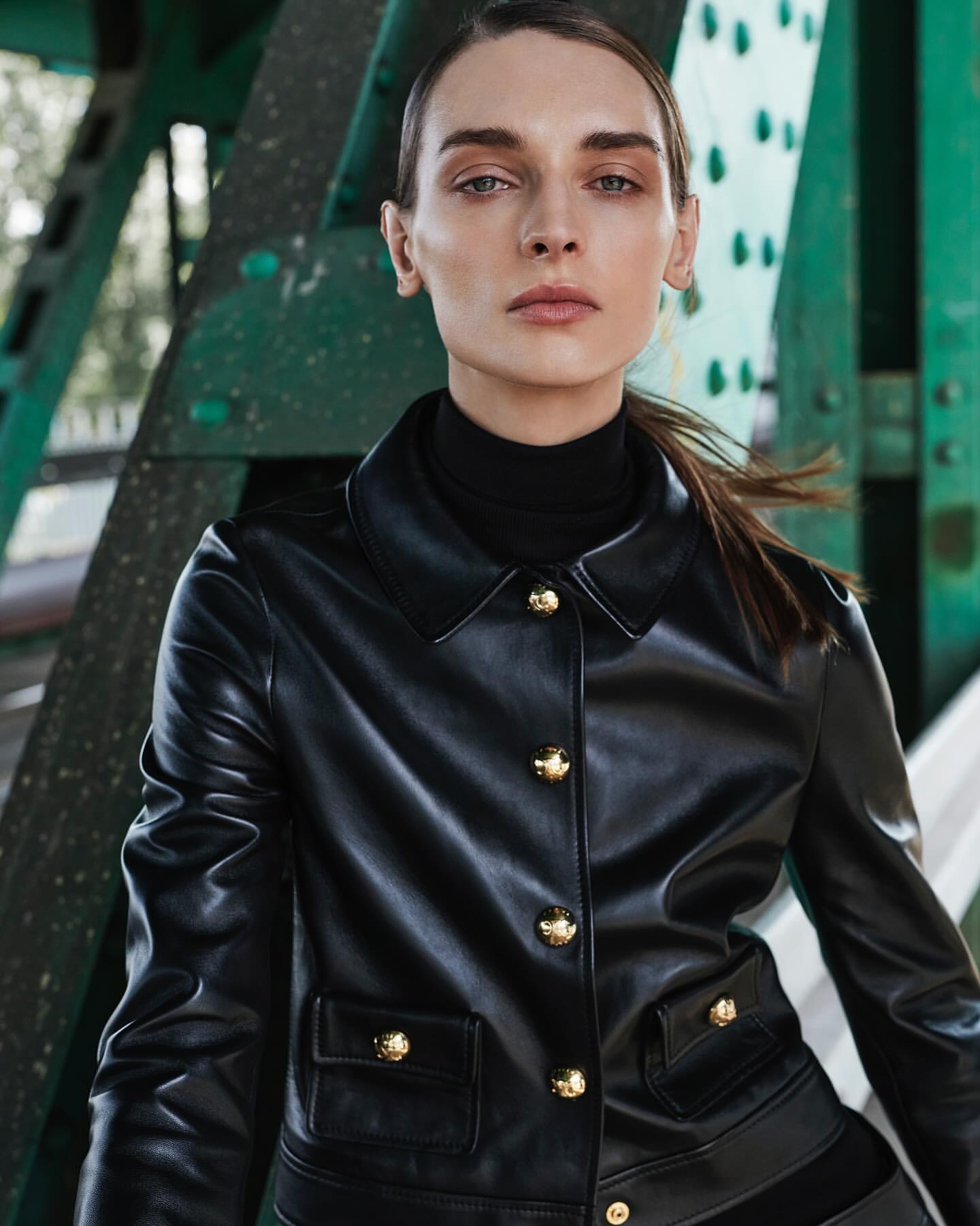 Portrait of a woman in a black jacket standing in front of a green metal structure, with a shallow depth of field. Fashion editorial.