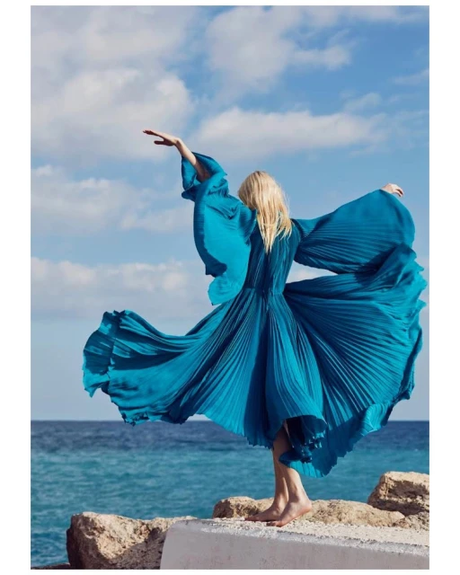 A woman in a long turquoise dress stands on rocks overlooking the ocean, with her arms raised against a partly cloudy sky.