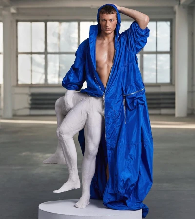 Man poses in oversized blue jacket with statue on a white plinth, inside an industrial-style space featuring large windows and concrete floor.
