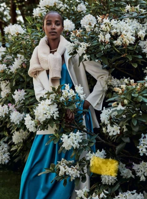 An adult female stands in a garden surrounded by white rhododendron flowers wearing a blue dress, cream jacket and sweater.