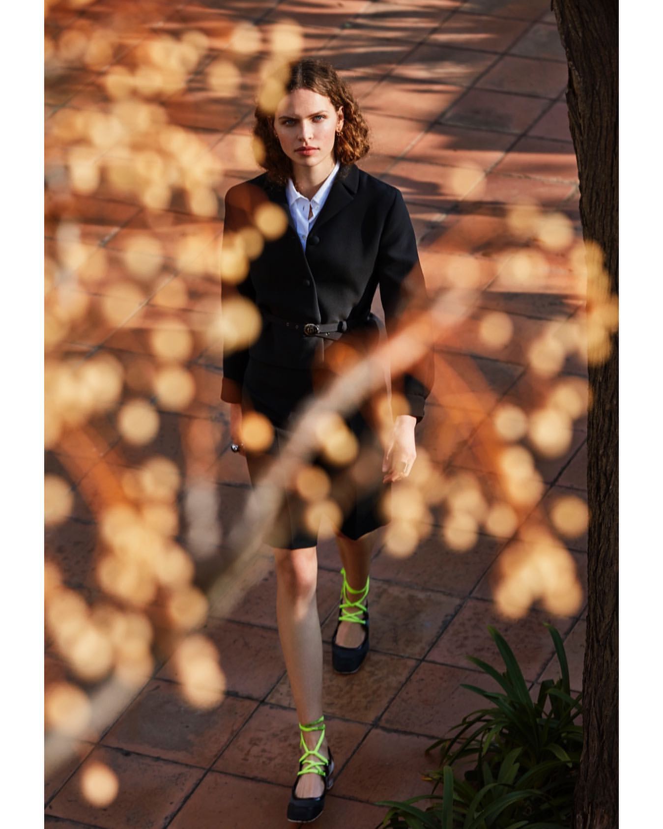 Fashionable adult woman walks on brick path outdoors, wearing a black blazer, skirt, and neon green strappy flats, with bokeh in foreground.