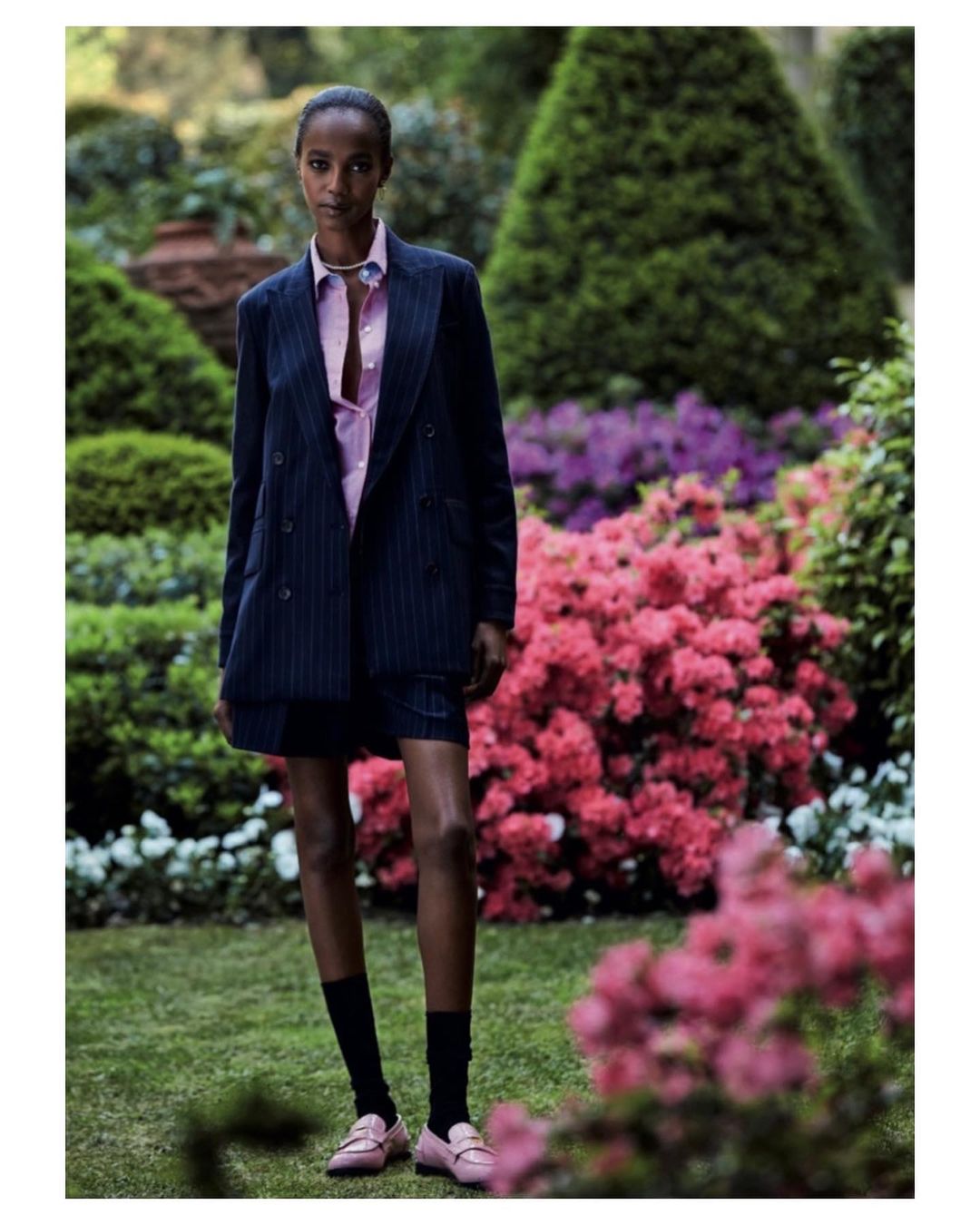 Fashion shot of a Black female model in a pinstripe suit, standing in a garden with pink flowers. She wears pink loafers and black socks.