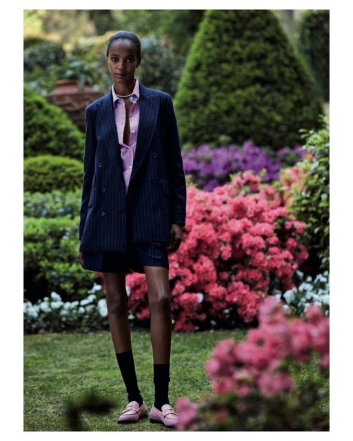 Fashion shot of a Black female model in a pinstripe suit, standing in a garden with pink flowers. She wears pink loafers and black socks.