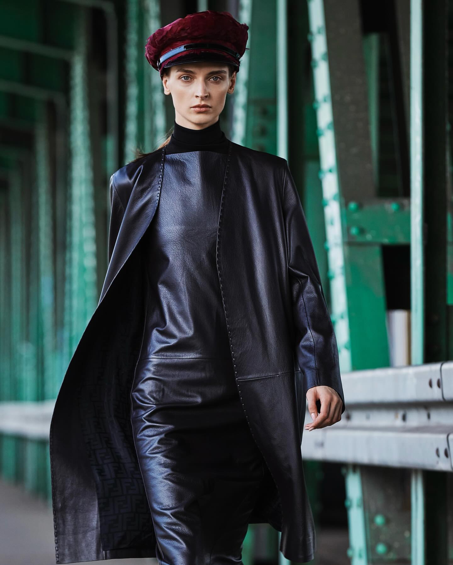 A stylish adult female strides on a green bridge wearing a black leather-like dress, matching duster coat, and a maroon beret.
