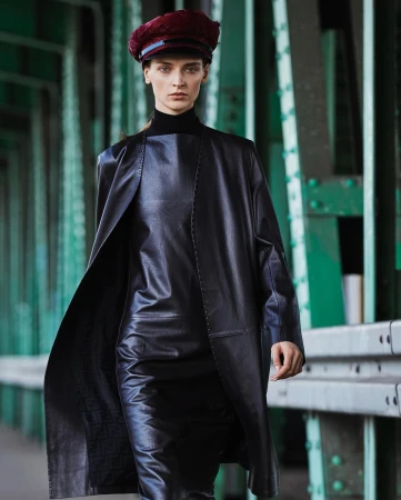 A stylish adult female strides on a green bridge wearing a black leather-like dress, matching duster coat, and a maroon beret.