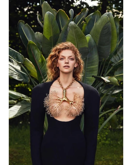 A curly-haired woman in a black dress poses against a lush green backdrop. The dress features a lung-shaped gold necklace.