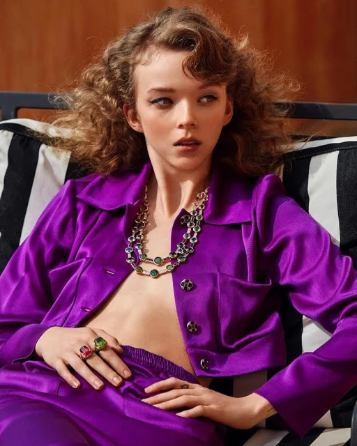 Portrait of a stylish young woman with curly hair wearing a purple satin suit and statement jewelry posing indoors, beauty and fashion shot.