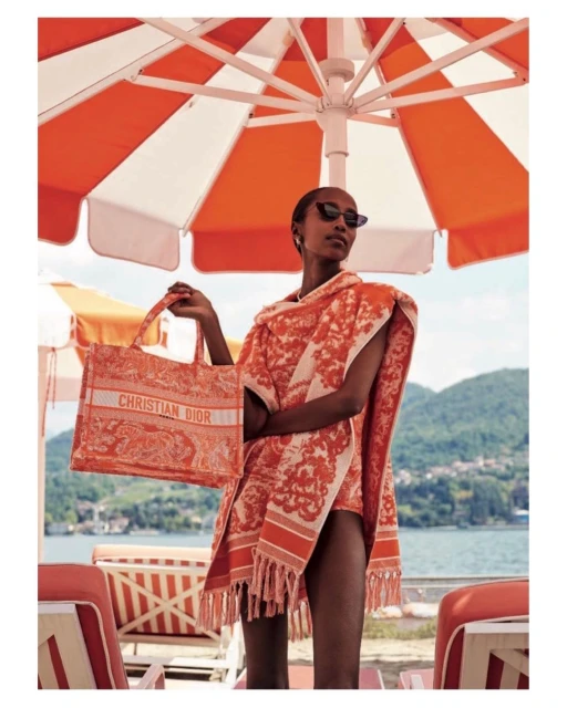 Woman at outdoor resort holds branded tote bag under orange and white umbrella, wearing printed cover-up, with a lake view.