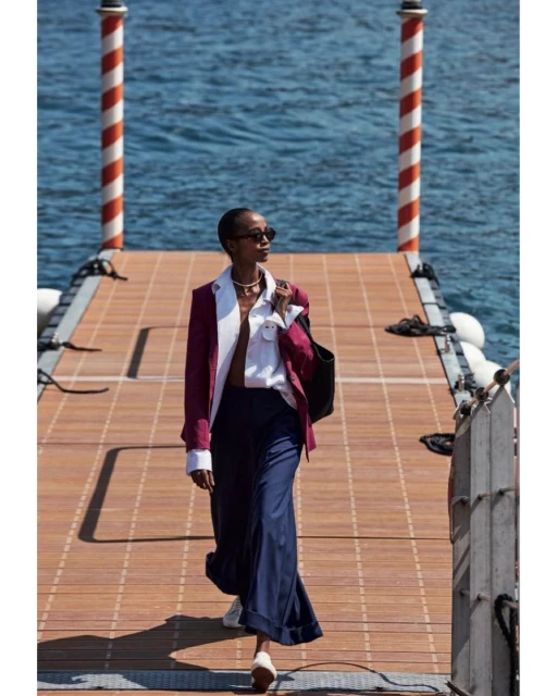 Woman in stylish jacket and wide-leg pants walks on a pier with the ocean in the background on a sunny day with bright lighting.