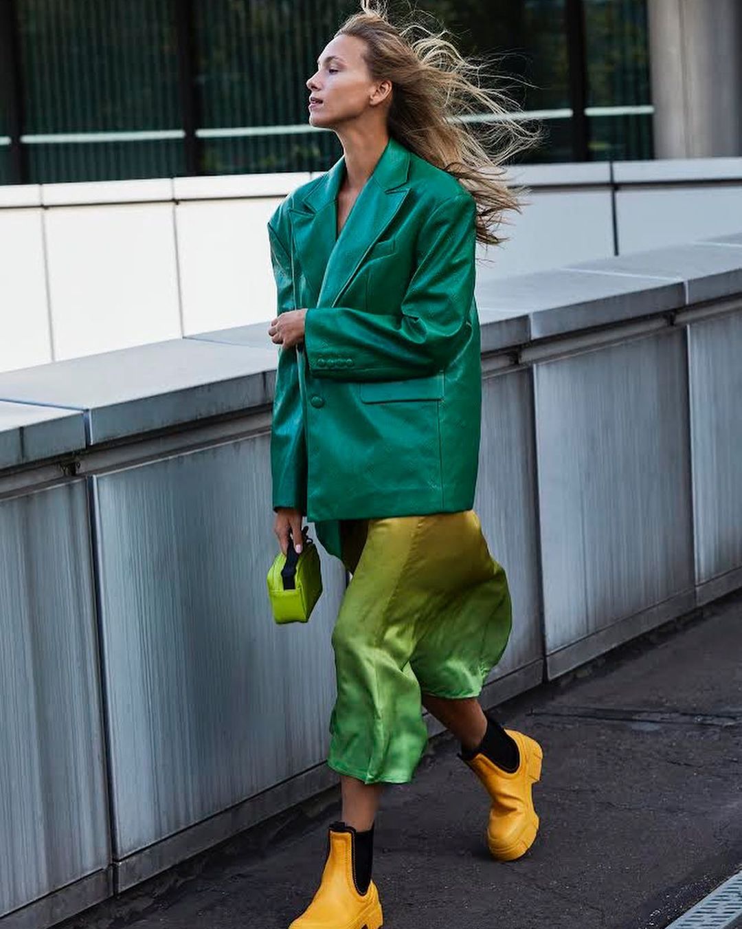 A woman walks outside wearing a green blazer, yellow and green skirt, and yellow boots, carrying a small green purse.