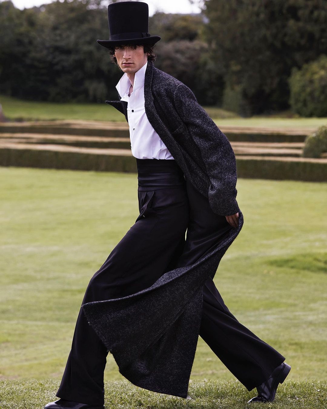 A person in a dark coat and top hat walking across a lawn, surrounded by greenery and trimmed hedges in the background, fashion.