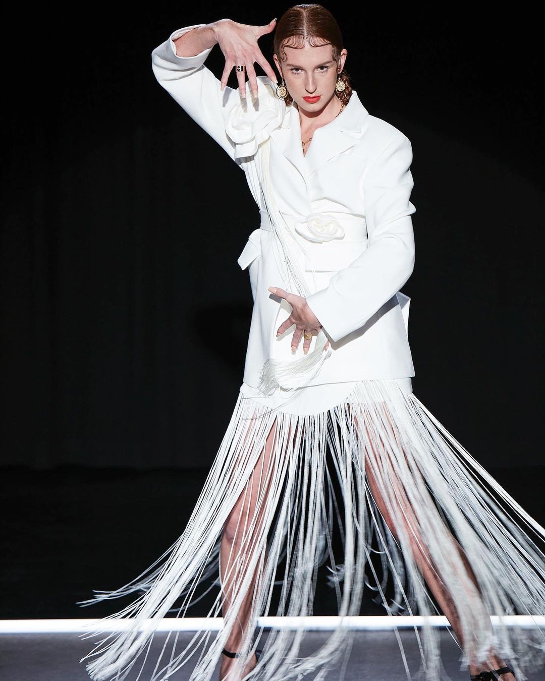 Elegant adult woman poses wearing a white jacket and fringe skirt against a dark background with studio lighting and outstretched hands.