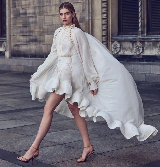 A woman in a long, white dress walks outdoors with a flowing cape. The dress has ruffles, long sleeves, and a high neckline.