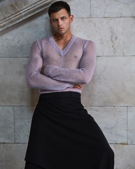 Portrait of an adult male with crossed arms wearing a see-through purple sweater and a black skirt leaning against a marble wall.