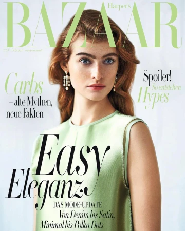 Portrait of a red-haired woman in a light green sleeveless top wearing pearl drop earrings against a light textured background