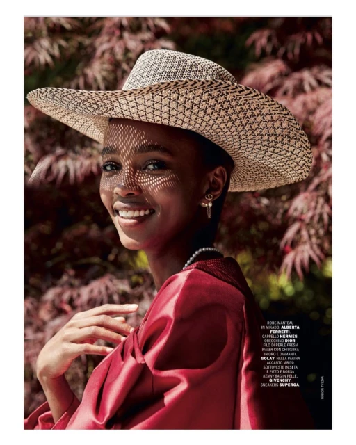 Close-up portrait of a smiling Black woman wearing a wide-brimmed hat, red dress, and pearl necklace, posing outdoors with greenery.