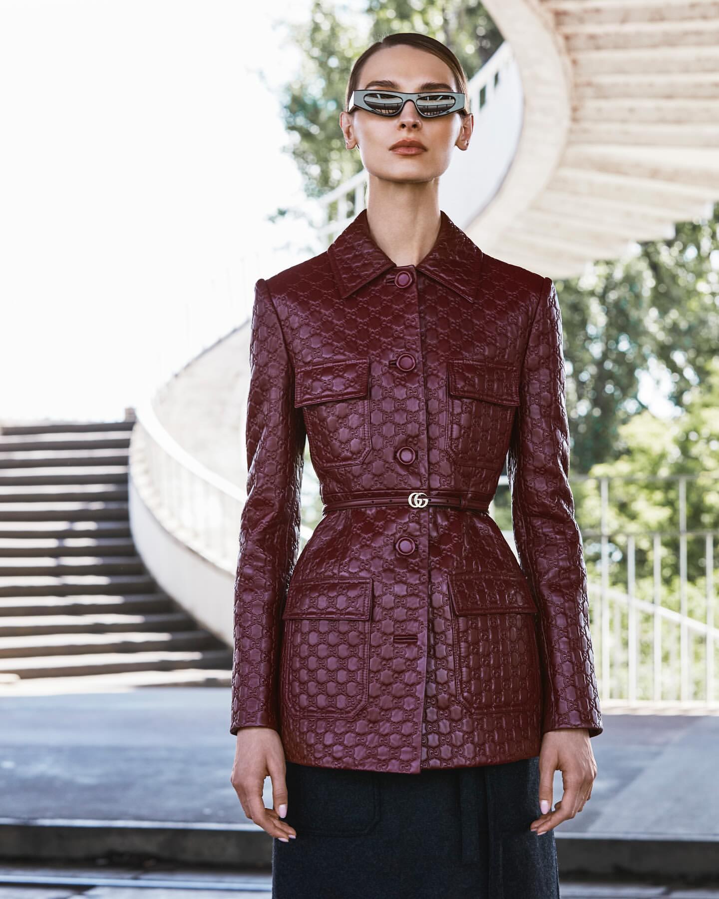 A female fashion model poses in a patterned burgundy leather jacket and stylish sunglasses in front of a spiral staircase and trees.