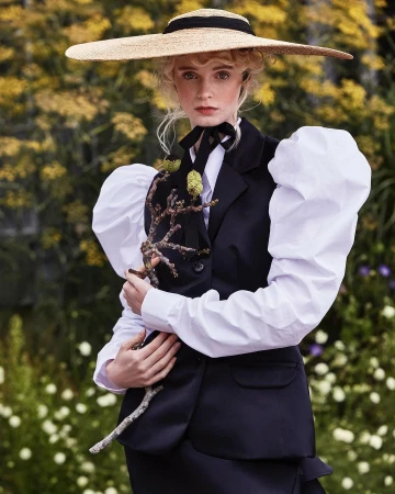Woman in black vest, white blouse, and straw hat holds a branch in a floral outdoor setting with flowers and greenery visible.