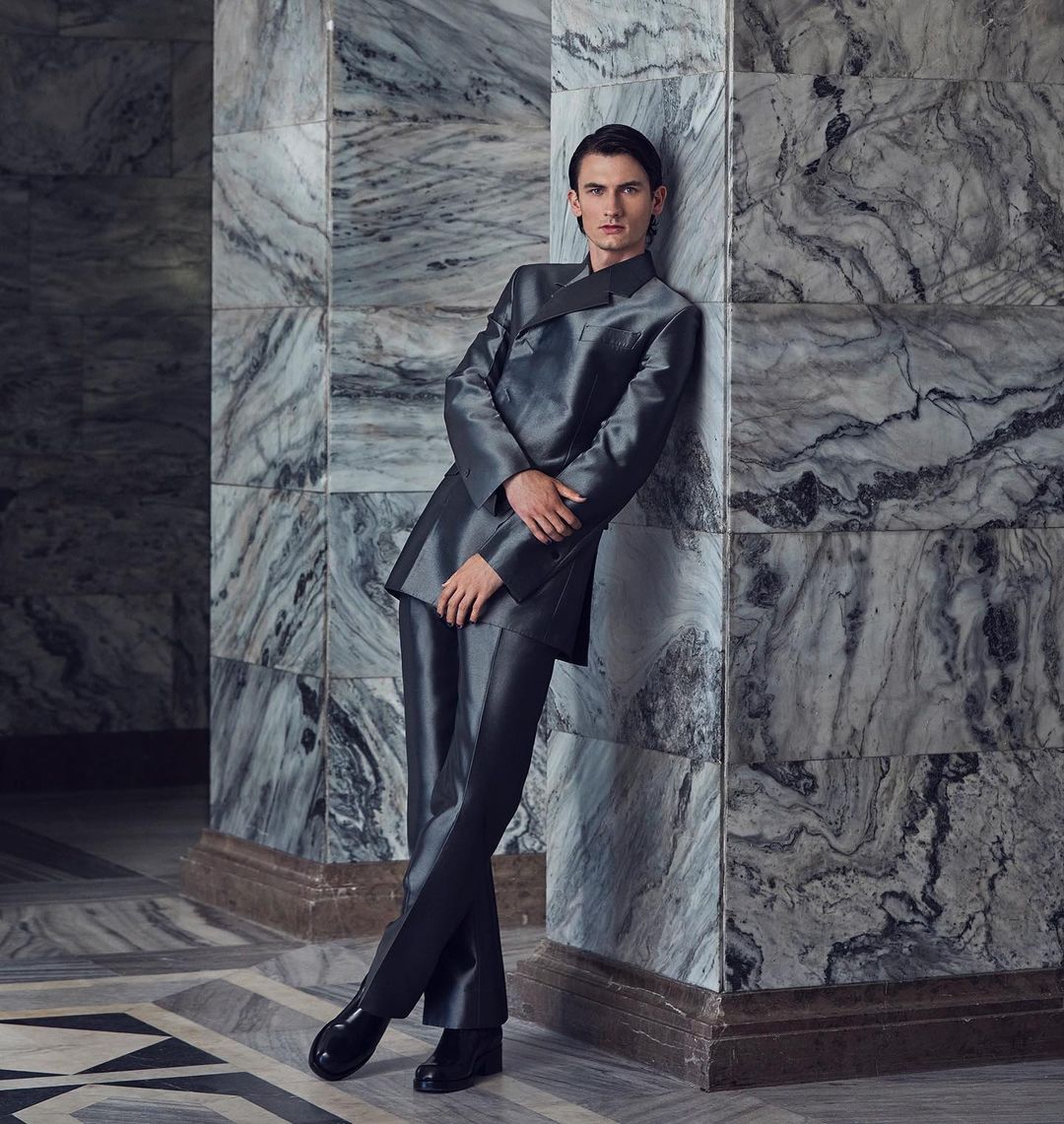 Young adult male posing in a shiny grey suit against a marble wall, captured in medium shot for a fashion or editorial aesthetic.