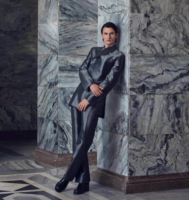 Young adult male posing in a shiny grey suit against a marble wall, captured in medium shot for a fashion or editorial aesthetic.