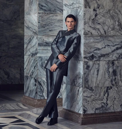 Young adult male posing in a shiny grey suit against a marble wall, captured in medium shot for a fashion or editorial aesthetic.