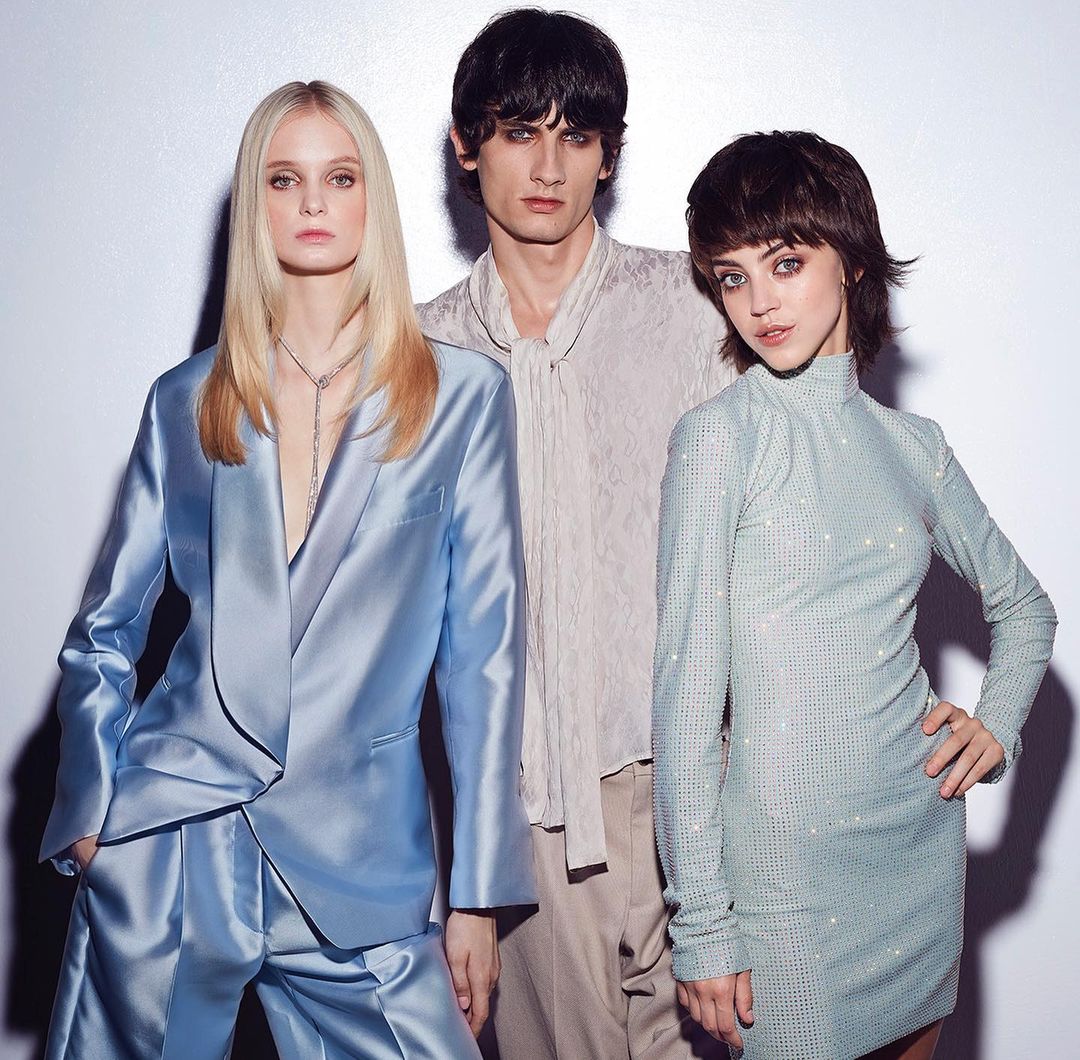 Three fashion models pose in a studio setting. Two women wear light-colored dresses and suits, while a man wears a light shirt.