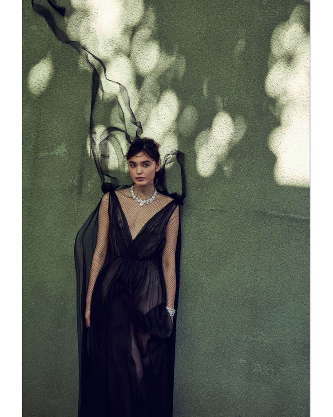 Woman in an elegant black gown stands against a textured green wall with dappled sunlight, exuding sophisticated fashion and style.