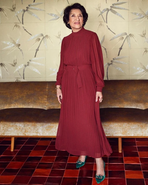 Full shot of a mature woman in a burgundy dress and green heels standing in front of a patterned wallpaper and brown couch.