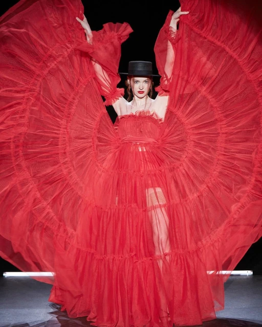 Elegant fashion photo of a woman in a flowing red gown and black hat, posing against a dark background with stage lighting. 