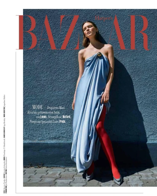 Editorial fashion shot of a woman in a draped dress and red tights standing against a textured blue wall with "BAZAAR" text overhead.