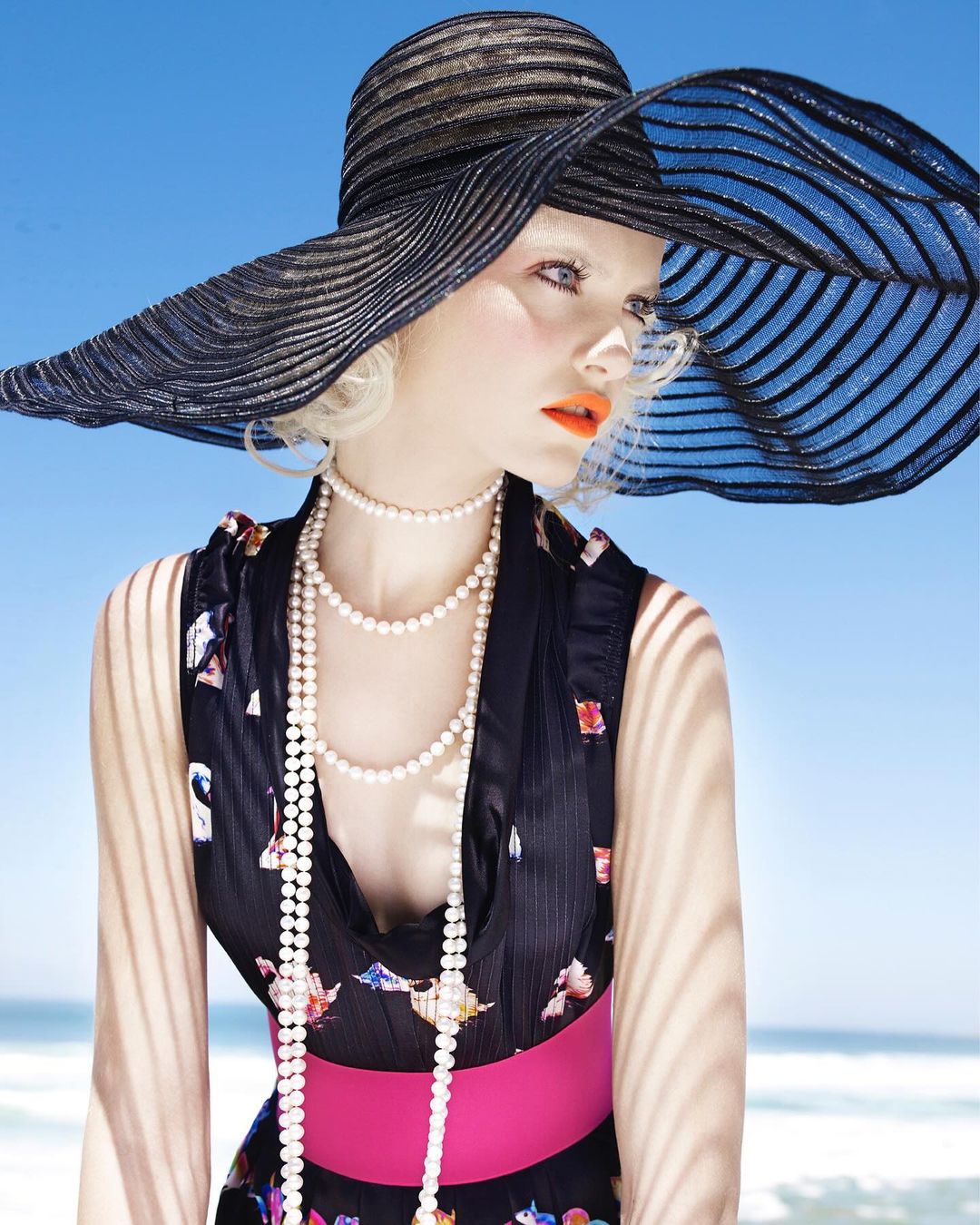 A fashion shot of a fair-skinned woman in a large black hat, pearl necklaces, and floral dress against a sunny beach backdrop.