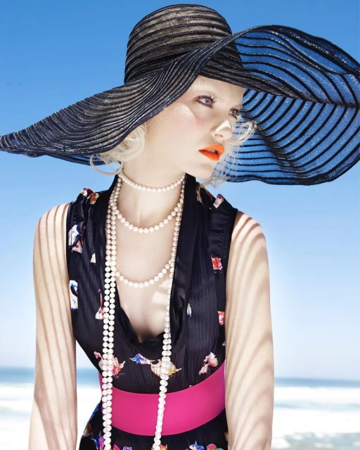 A fashion shot of a fair-skinned woman in a large black hat, pearl necklaces, and floral dress against a sunny beach backdrop.