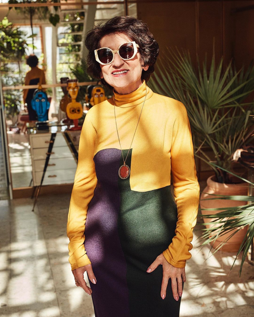 Attractive mature woman with sunglasses poses in a colorful dress inside a sunlit room filled with indoor plants and modern art.