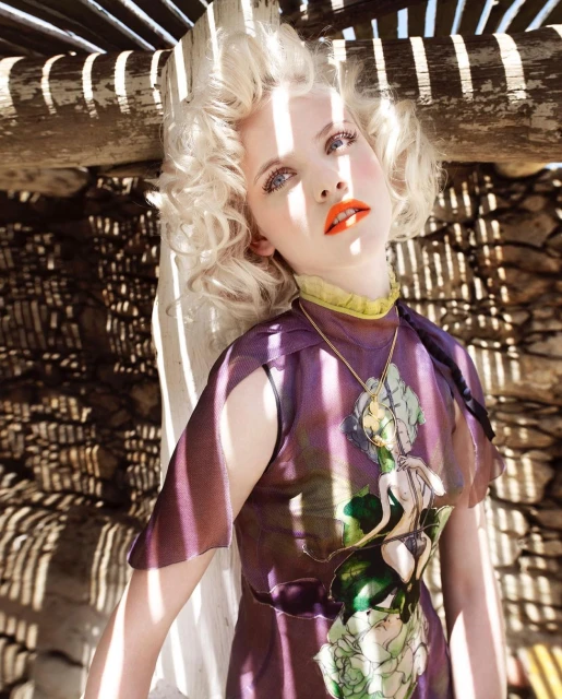 Elegant woman with blonde curly hair, wearing a purple dress with a graphic, in an outdoor setting with patterned shadows. 