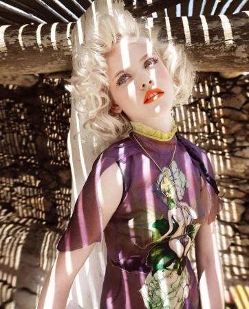Elegant woman with blonde curly hair, wearing a purple dress with a graphic, in an outdoor setting with patterned shadows. 