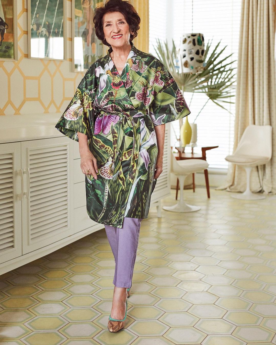Portrait of a smiling senior woman in a floral print robe and purple pants, standing in a sunlit living room interior with white furniture.