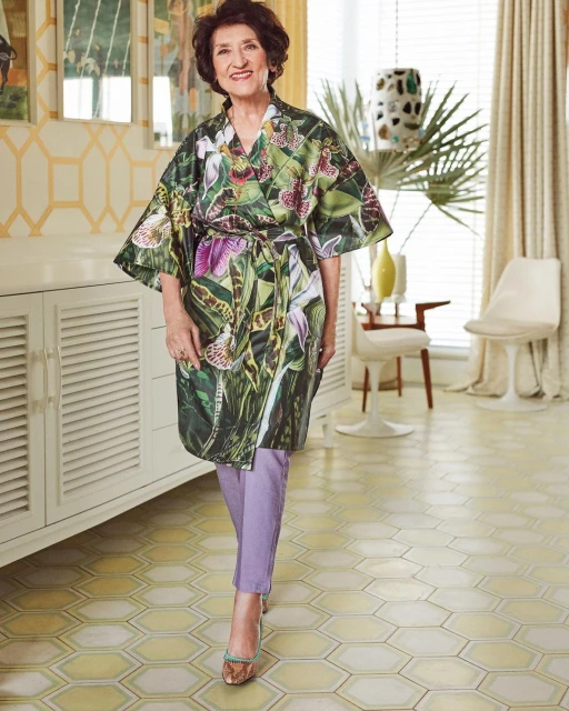 Portrait of a smiling senior woman in a floral print robe and purple pants, standing in a sunlit living room interior with white furniture.