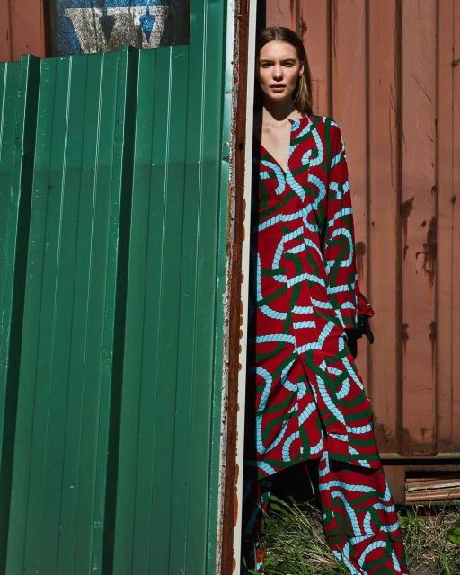 A fair-skinned young woman poses in a red and blue patterned top and pants set near a weathered green and brown corrugated metal wall.
