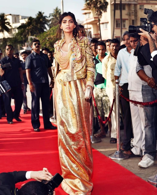 Fashionable woman in a gold ensemble poses confidently on a red carpet surrounded by photographers and onlookers outdoors.