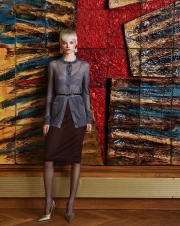 Model with blonde pixie cut wearing a sheer grey cardigan over brown pencil skirt standing against a textured painting.