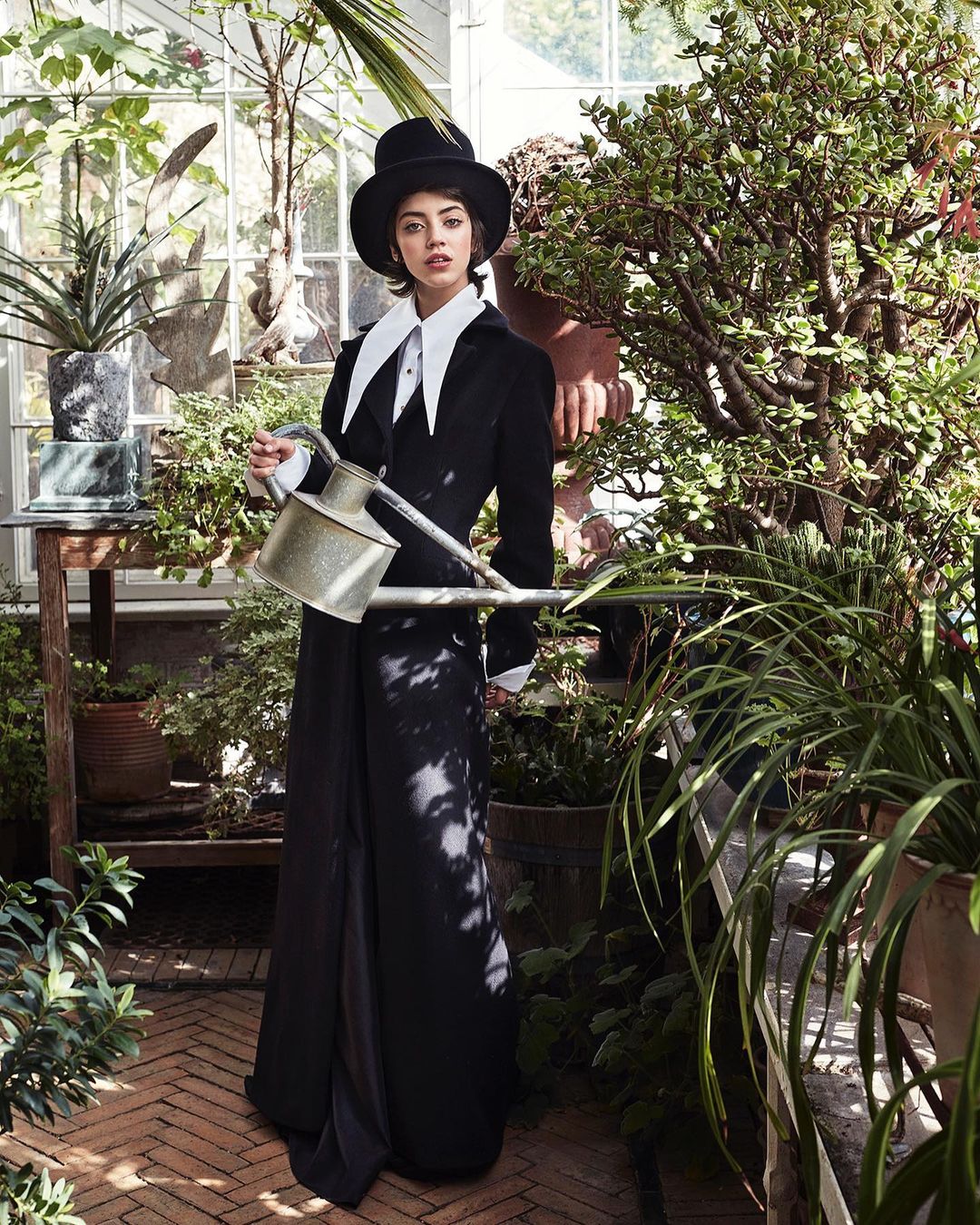 Woman in black coat and top hat holds watering can in a lush greenhouse, surrounded by vibrant green plants and flowers, stylish fashion.