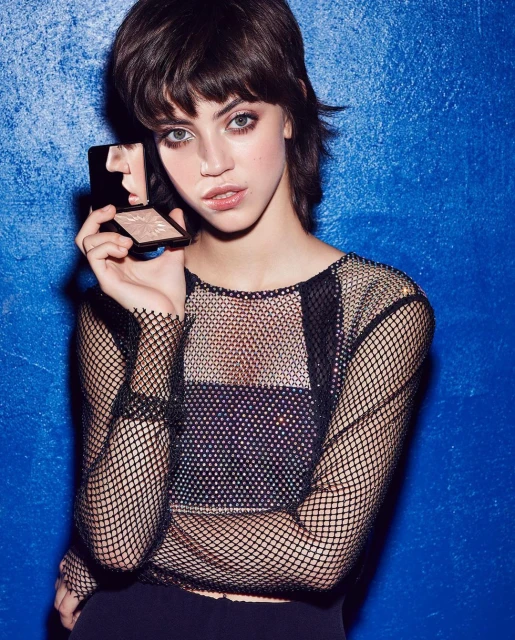 Young woman with short hair and fishnet top holds an open makeup compact in front of a textured blue background.