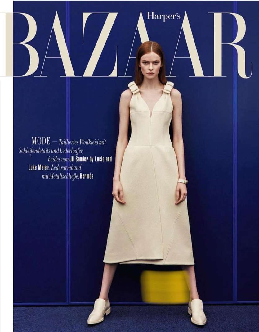 Fashion editorial shot of a model wearing a cream-colored dress and shoes, set against a blue backdrop with 'Harper's Bazaar' text overlay.