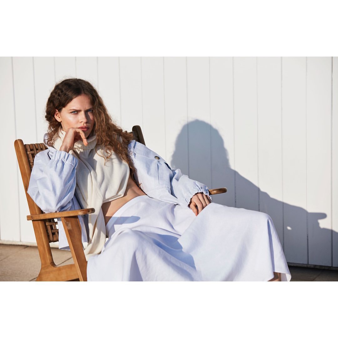 An adult woman with long brown hair sits in a wooden chair wearing a light blue jacket and a long white skirt in a relaxed pose.