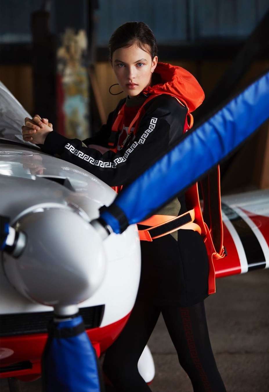 A brunette woman in a black and orange jacket leans against a small airplane with a red and white body and blue propeller in an aircraft hangar.