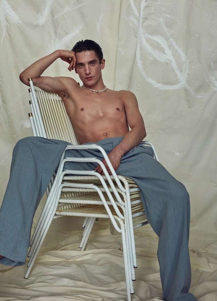 A young adult male sits shirtless in blue pants on a white chair with his hand on his head in a studio setting.