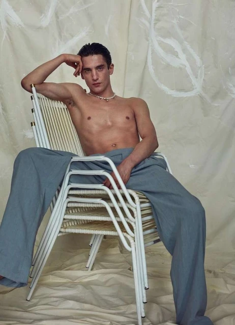A young adult male sits shirtless in blue pants on a white chair with his hand on his head in a studio setting.