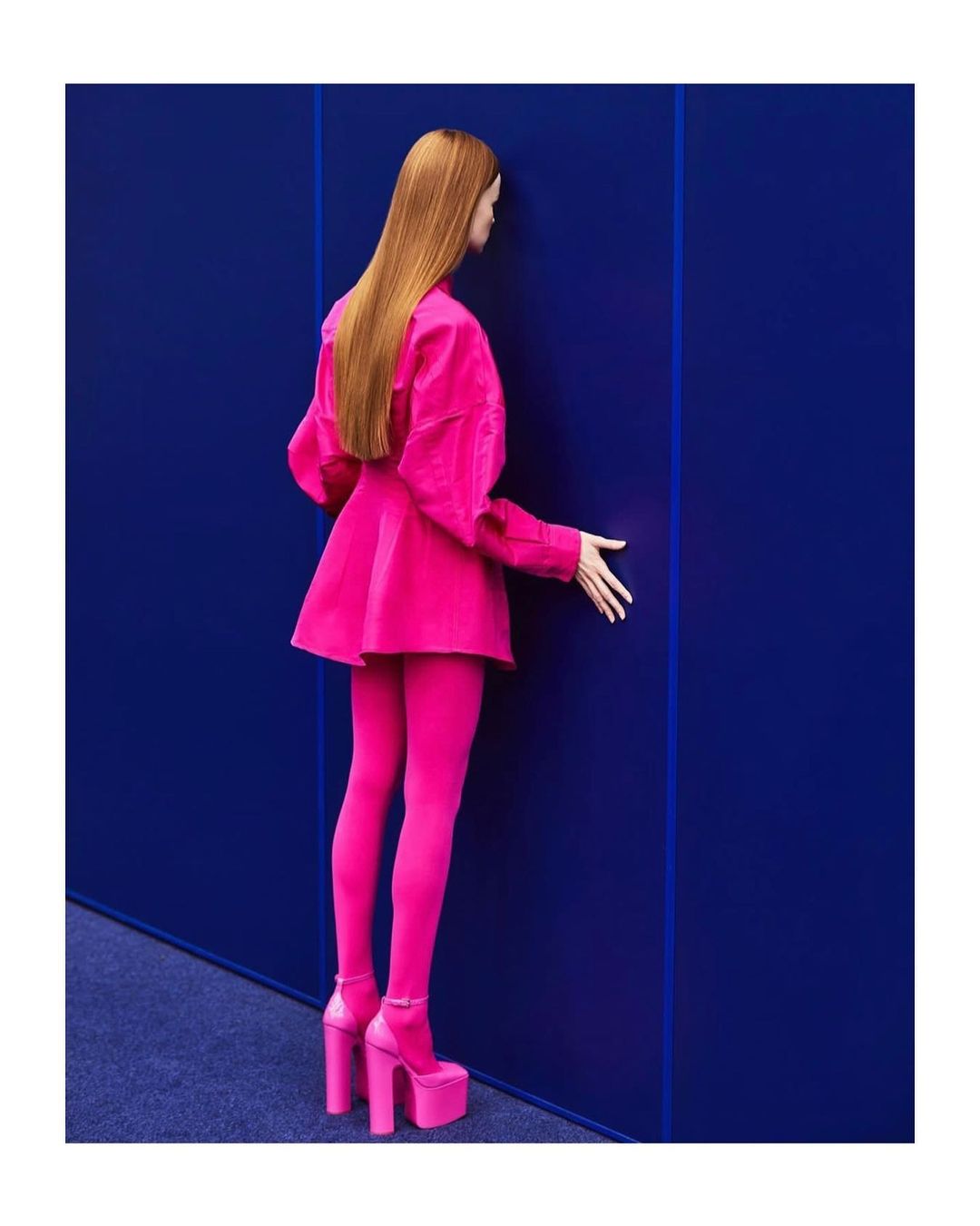Woman in bright pink skirt suit and platform heels poses against a royal blue wall in a minimalist studio setting. Fashion photography.